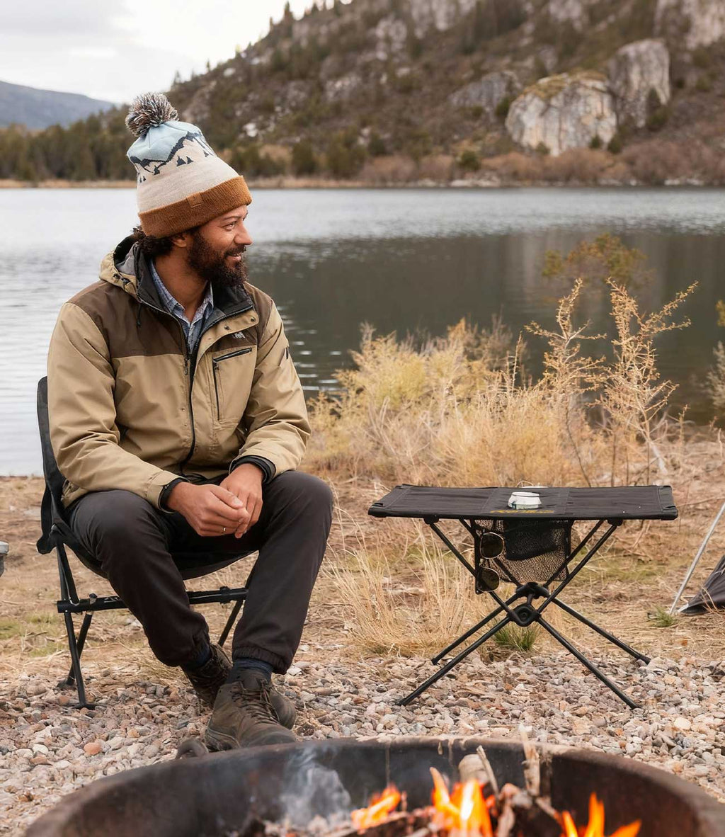 Man relaxing by campfire and lake with RoosCamp Portable Camping Table beside him in an autumn mountain setting