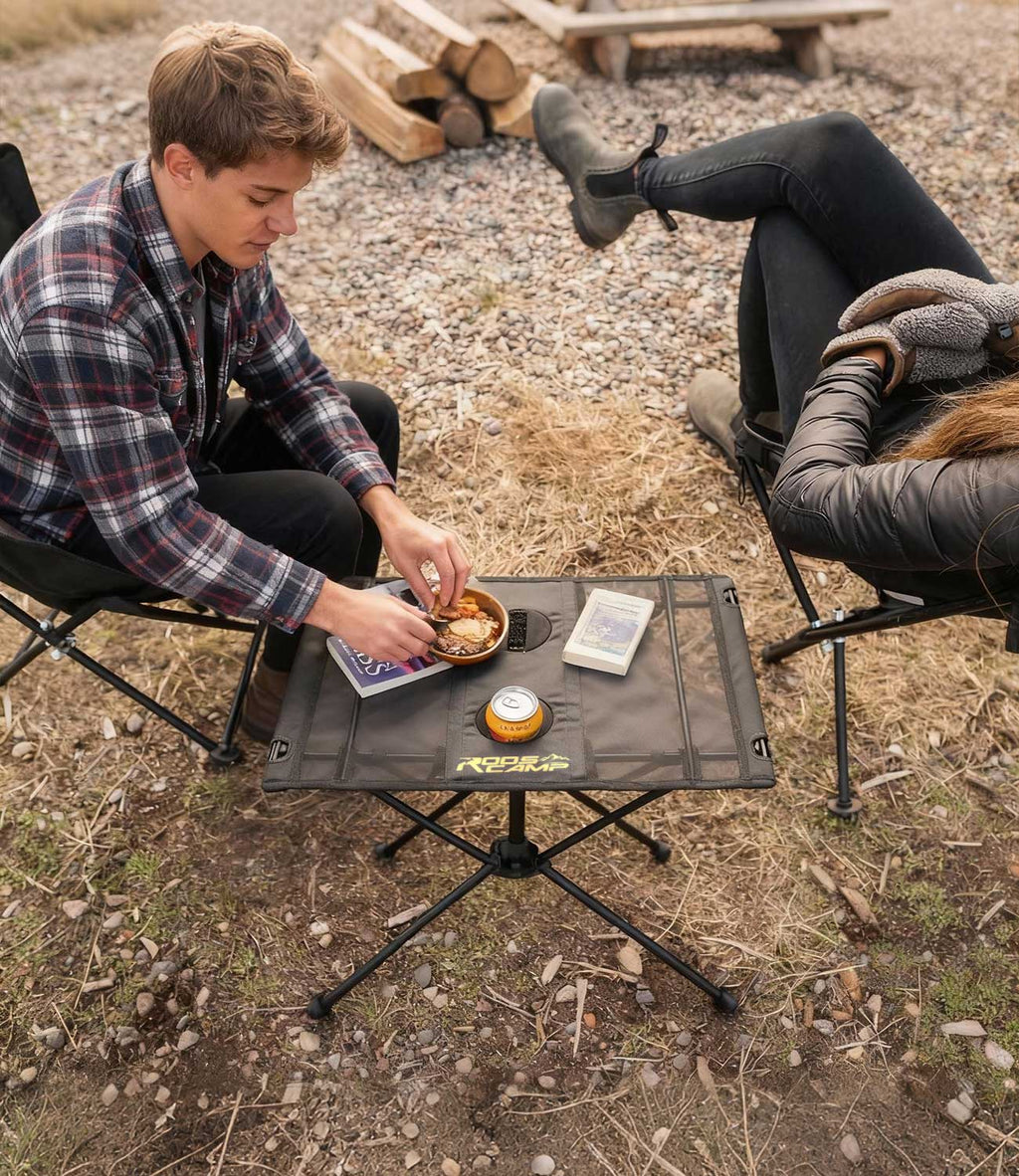 Couple at campsite using RoosCamp Portable Camping Table with food, drink and book on the surface, viewed from above