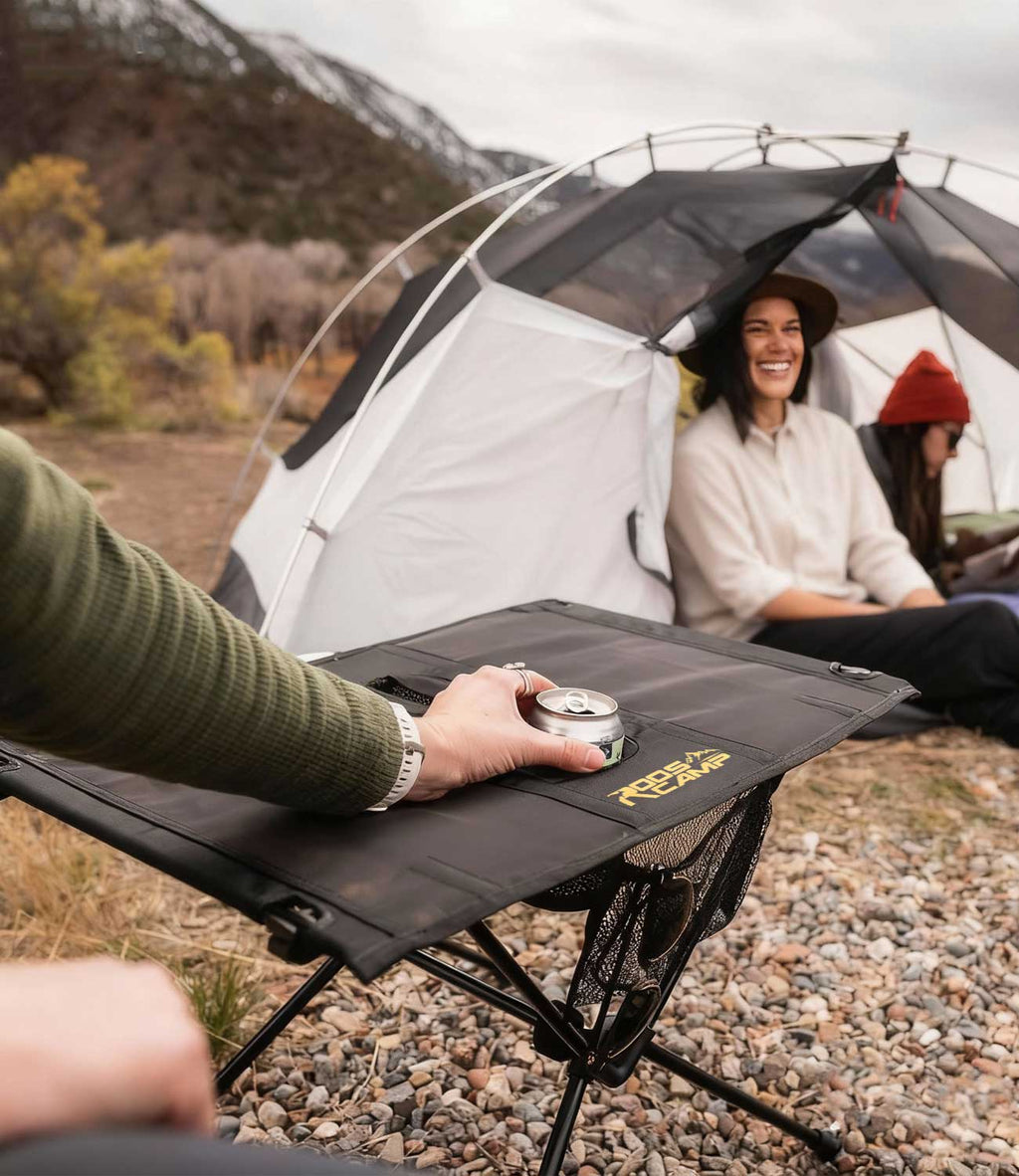 Friends camping in mountains with RoosCamp Portable Camping Table in foreground, drink can in cup holder, tent and laughing woman in background